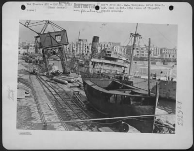 Thumbnail for Consolidated > East side of Pier E shows 380-foot merchant vessel (1) aground. Direct bomb hit on edge of pier has ripped up railroad track, up-ended a crane. Demolition of many sunken vessels, to clear docks, was hampered by ammunition holds.