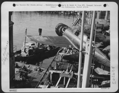 Thumbnail for Consolidated > "Off we go . . ." Not into "the wild blue yonder," but over the side of the Liberty ship. Still a helpless creature, it can fly only as high as the crane operator wills.