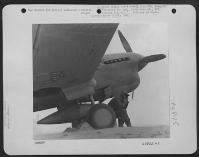 Thumbnail for Consolidated > A fighter pilot of the S.A.A.F. huddles in the shelter of his aircraft as a sand storm sweeps over a landing ground in Libya. He has turned his collar up over his ears to protect his face from the sand.