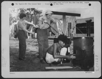 Consolidated > It's Monday (washday) at a WAC camp somewhere in New Guinea. T/4 Gillie Tanksley of Bokoshe, Oklahoma, hangs up some of the wash while T/4 Berta Hodnett of Notasulga, Alabama, lifts clothes out of the boiler. A native assists in building the fire.